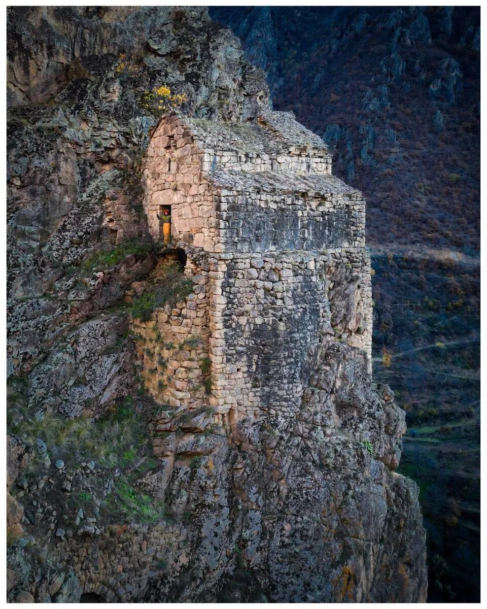 Nuka Sakdari seen from the air: a stone basilica standing on a narrow ledge of rock high above a wooded valley, its roof open to the sky.