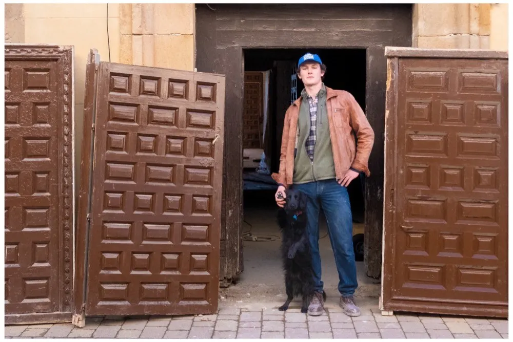 Four tall panelled wooden doors standing upright on the street outside a doorway, a man in a cap and leather jacket between them with a black dog at his feet