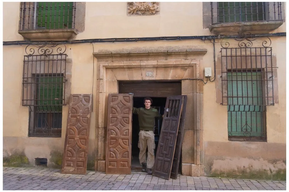 Ochre palace facade with iron-grilled windows, a carved wooden door leaning against the opening and a man standing in the dark doorway