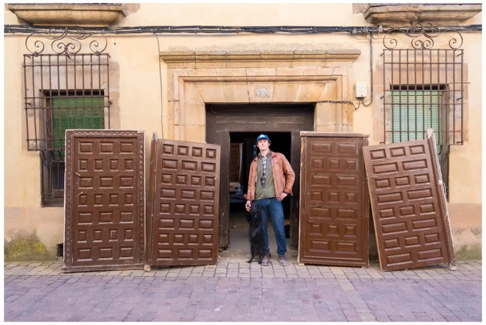 A row of heavy panelled wooden doors leaning against a historic ochre facade, a man standing in the empty doorway behind them