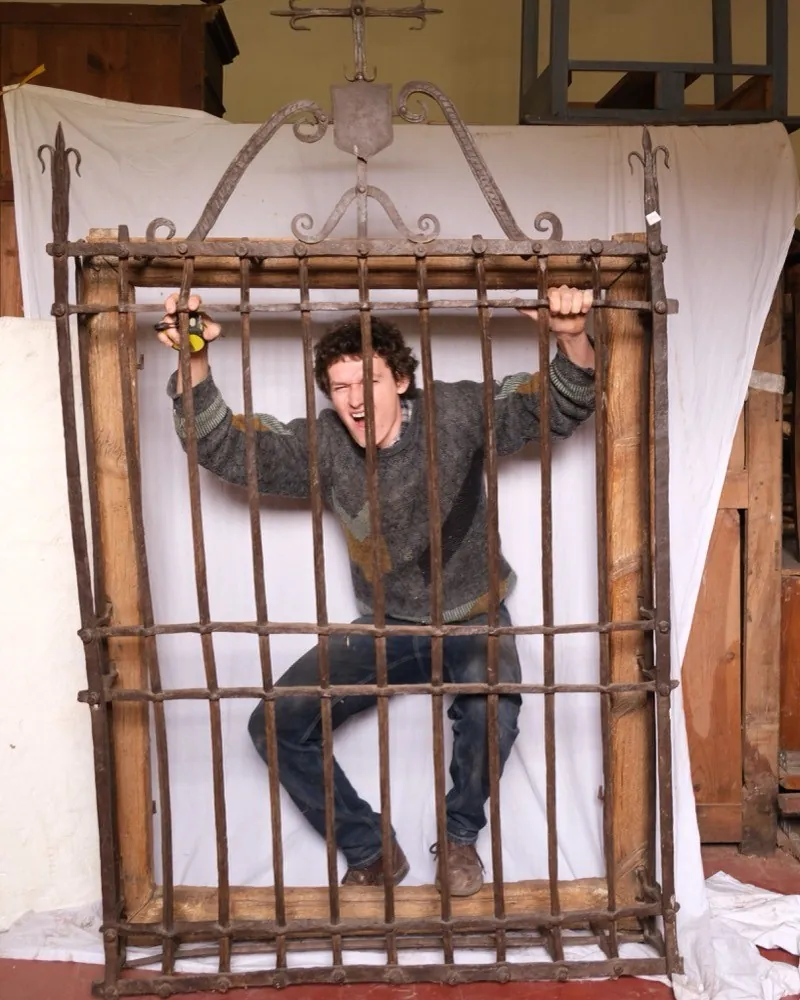 A man gripping the bars of a large wrought iron window grille mounted upright on a frame, pretending to be caged behind it
