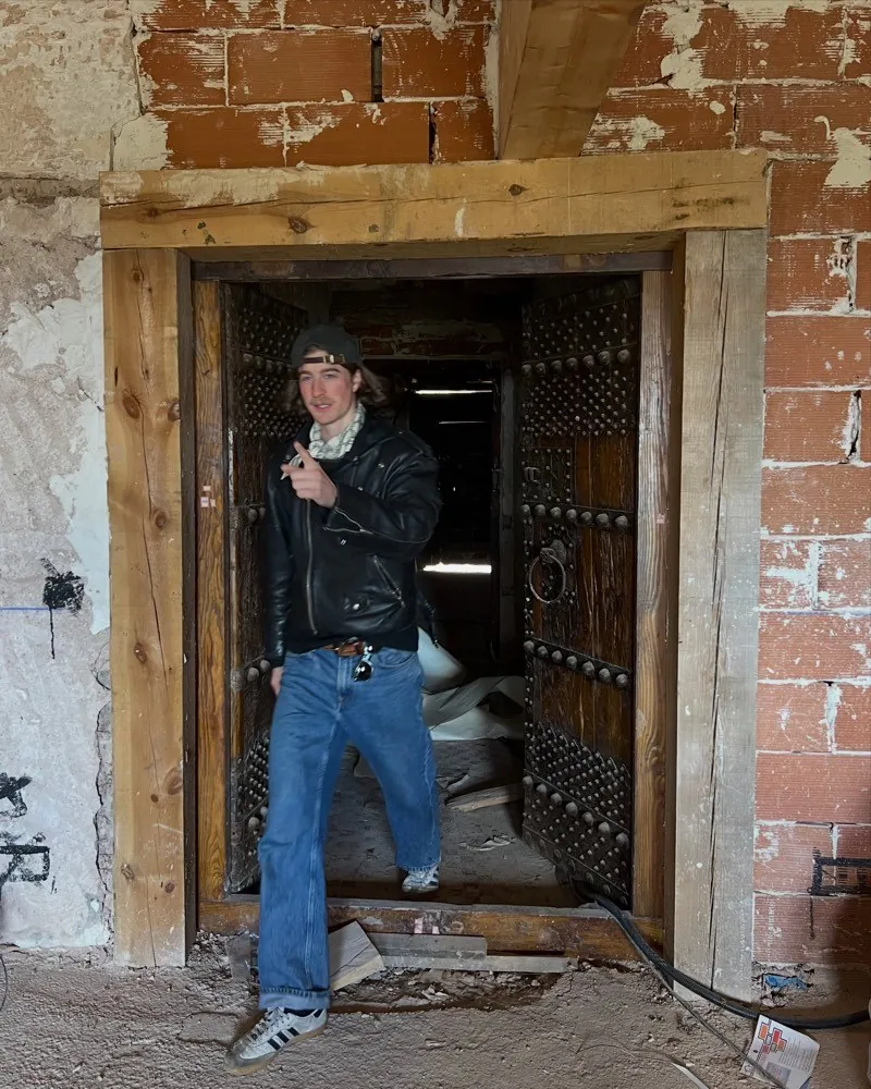 A man standing in the opening of an old studded wooden door in a stripped building with bare plaster and a raw timber lintel
