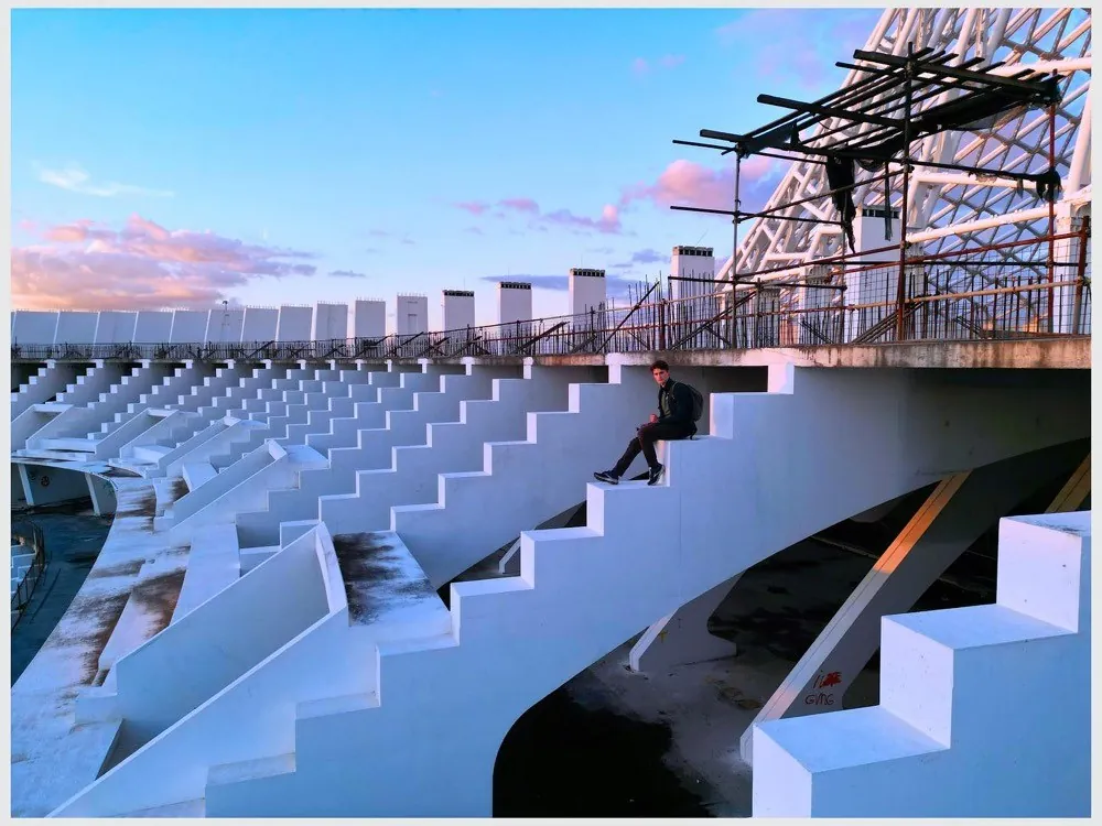 White concrete stadium steps rising towards scaffolding against a pink and blue dusk sky, a lone figure sitting on the edge
