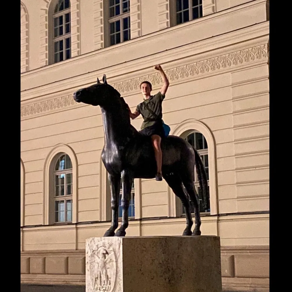 A bronze equestrian statue in front of a neoclassical facade, a young man sitting on the horse with one arm raised