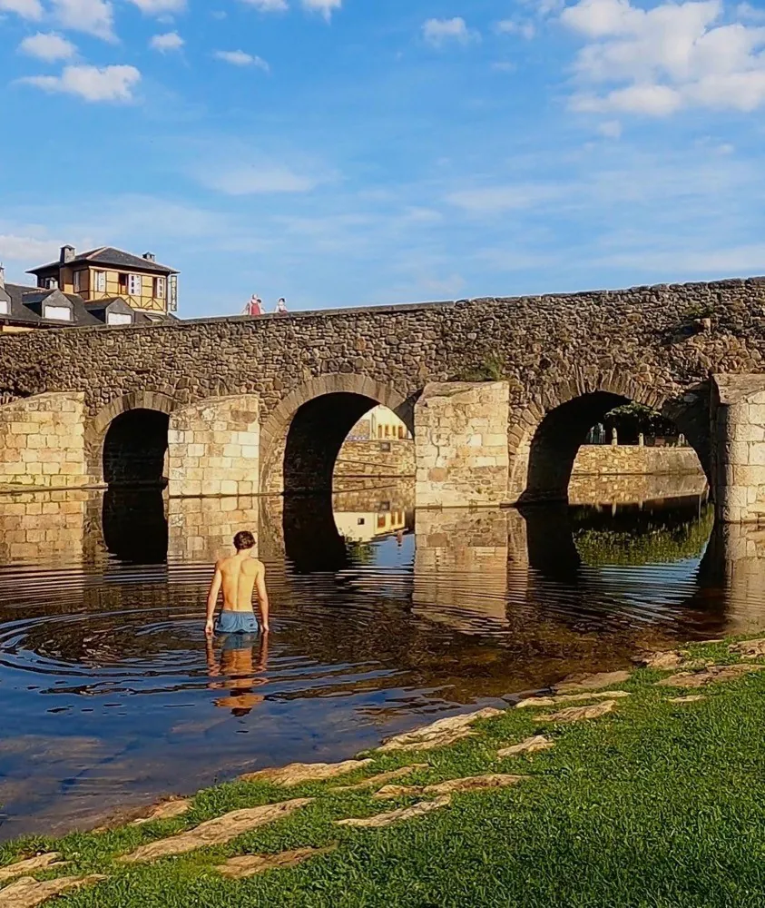 A Roman stone bridge of low arches over a calm river, a man standing waist deep in the water below it