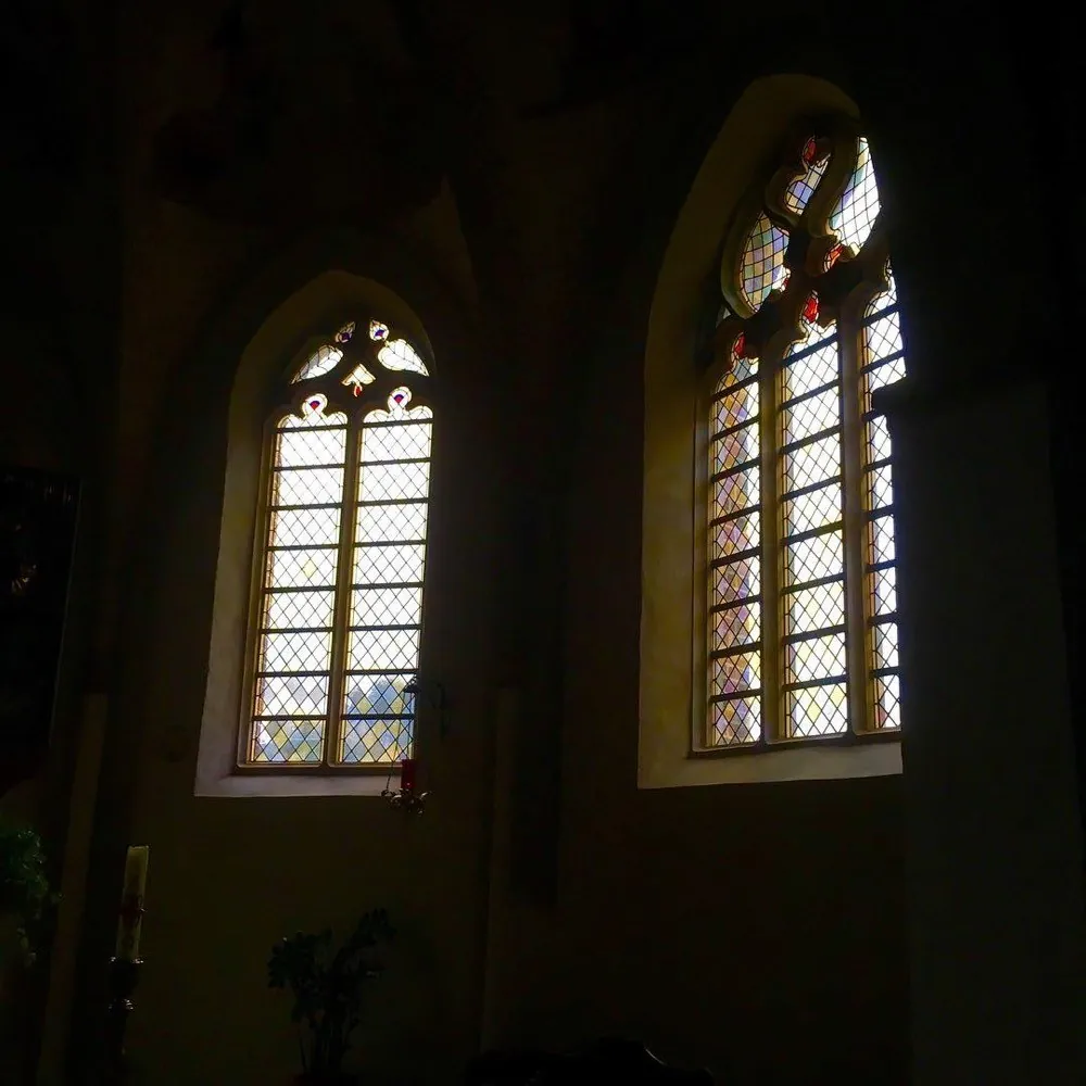 The dark interior of a church with two tall gothic windows of leaded glass glowing against black stone walls