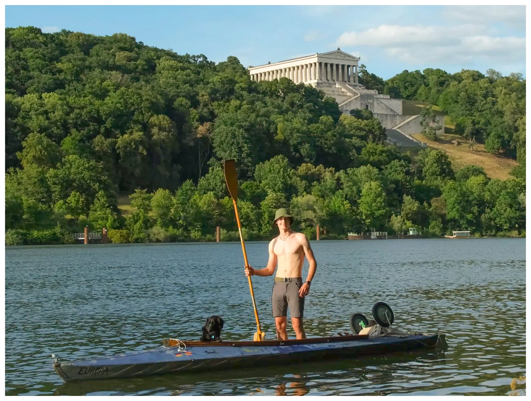 Tassilo standing upright on his loaded folding kayak, paddle raised, gliding a mirror-calm Danube at golden hour with Fluffo at the bow and the neoclassical Walhalla temple on the wooded hill behind
