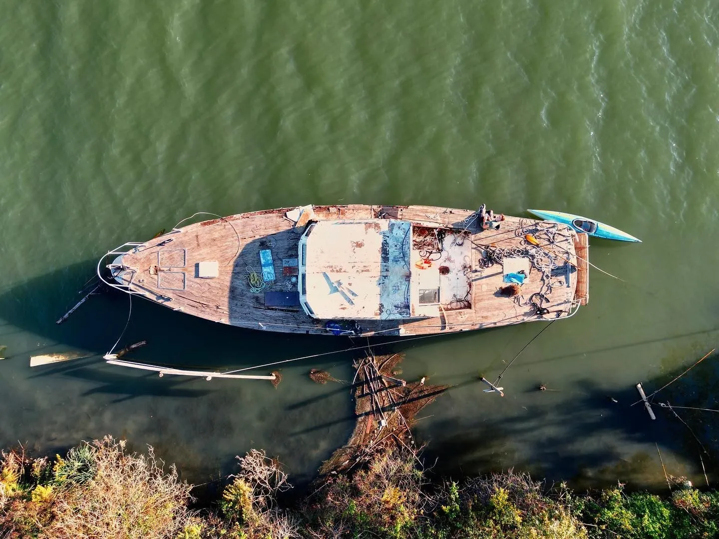 Drone view straight down onto a weathered wooden boat moored at a green riverbank, Tassilo's folding kayak tied alongside