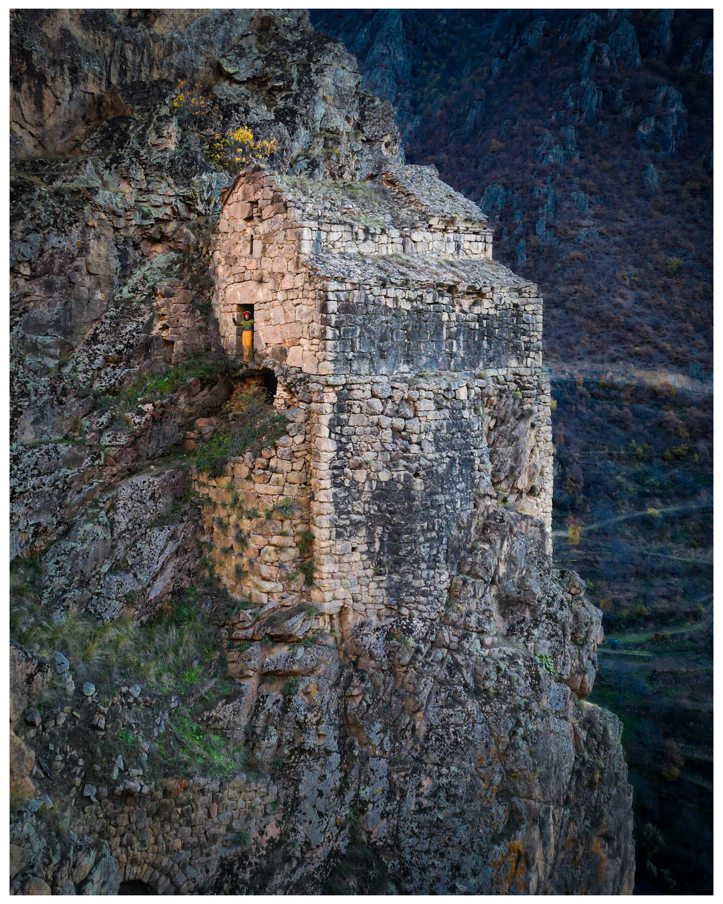 A ruined stone tower clinging to a sheer dark cliff face, a tiny figure in a red hat waving from its doorway