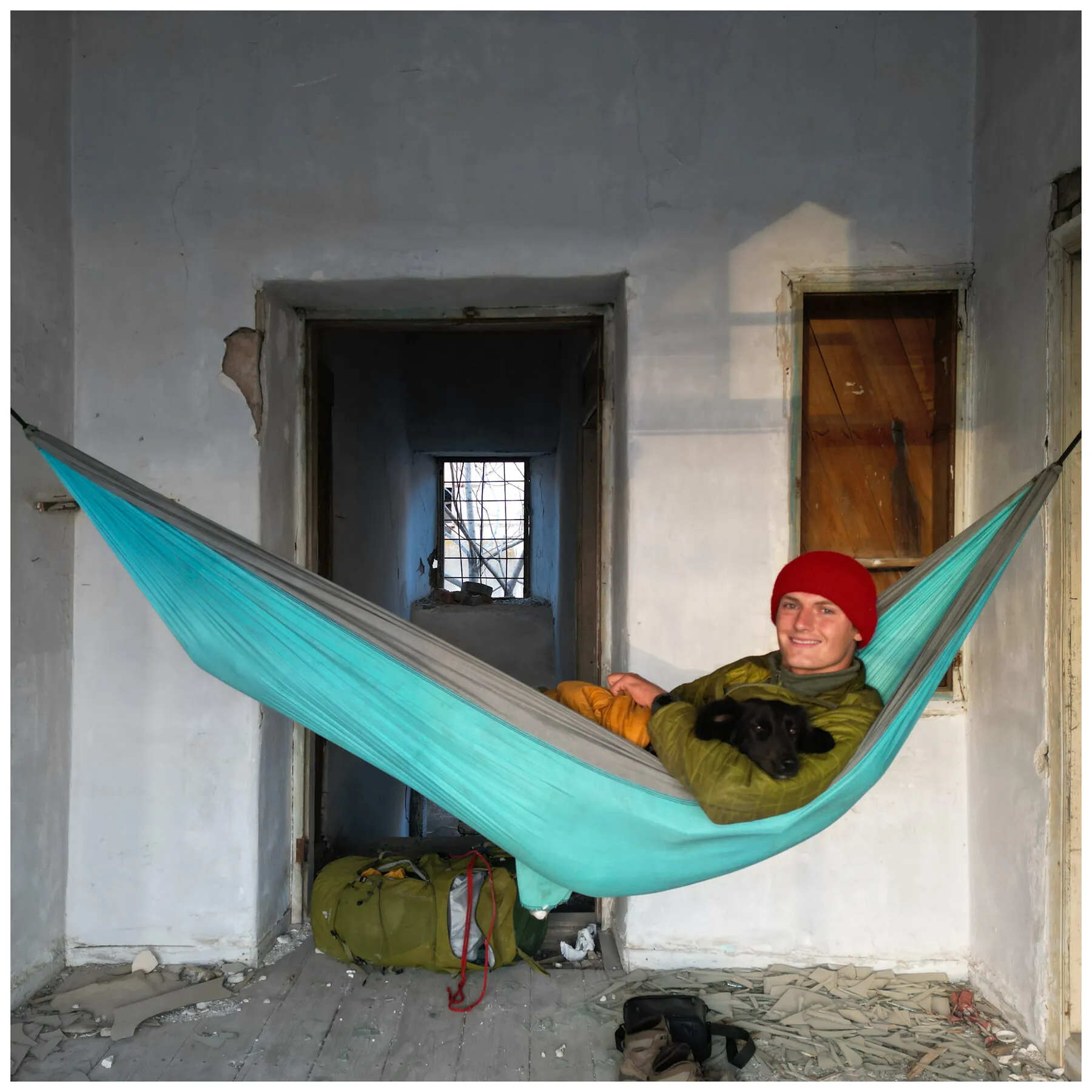 A teal hammock strung across a doorway in a derelict plastered room; Tassilo in a red wool hat rests inside it with Fluffo the black dog on his chest