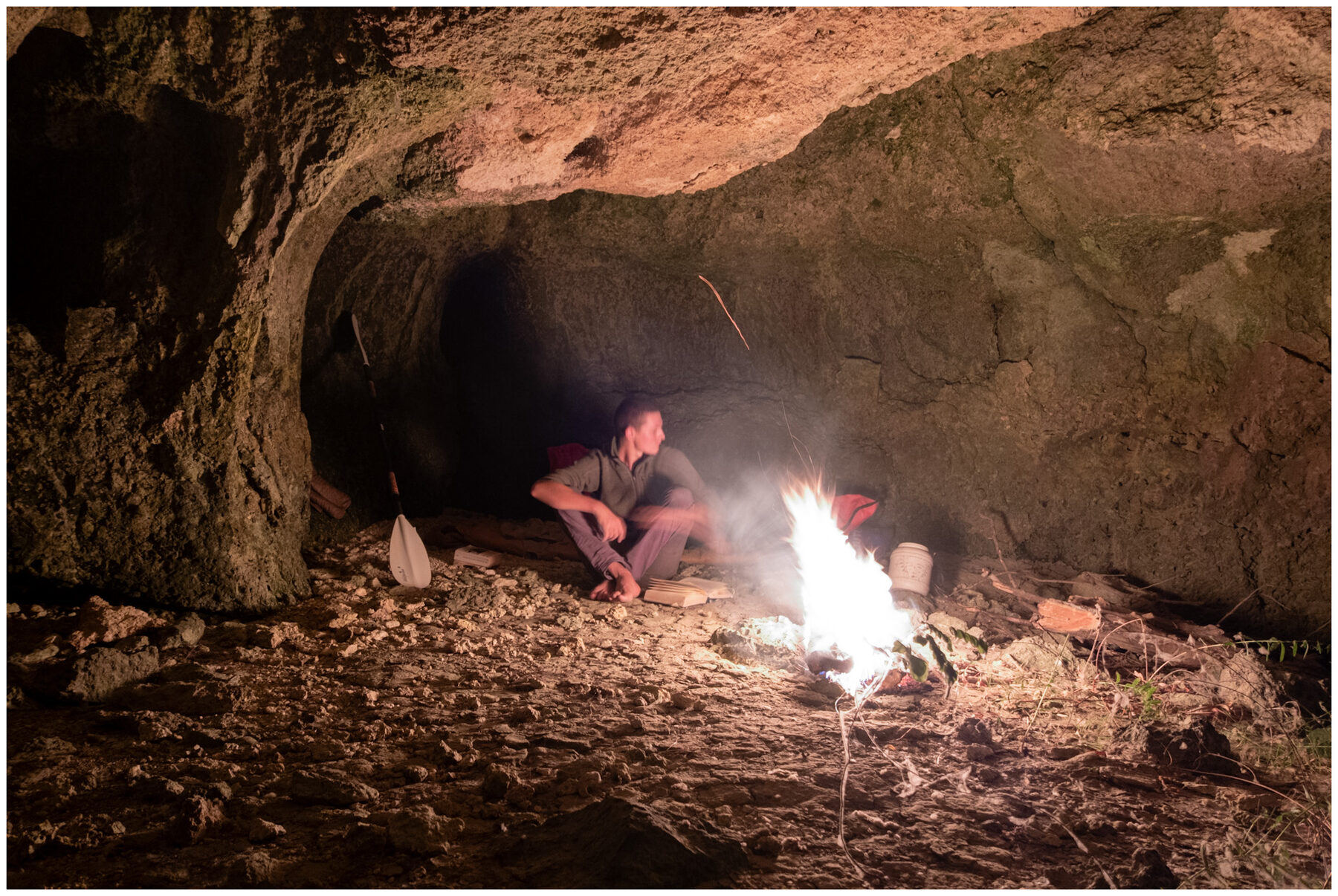 Tassilo sitting barefoot beside a bright campfire inside a dark rock cave at night, his kayak paddle leaning against the wall