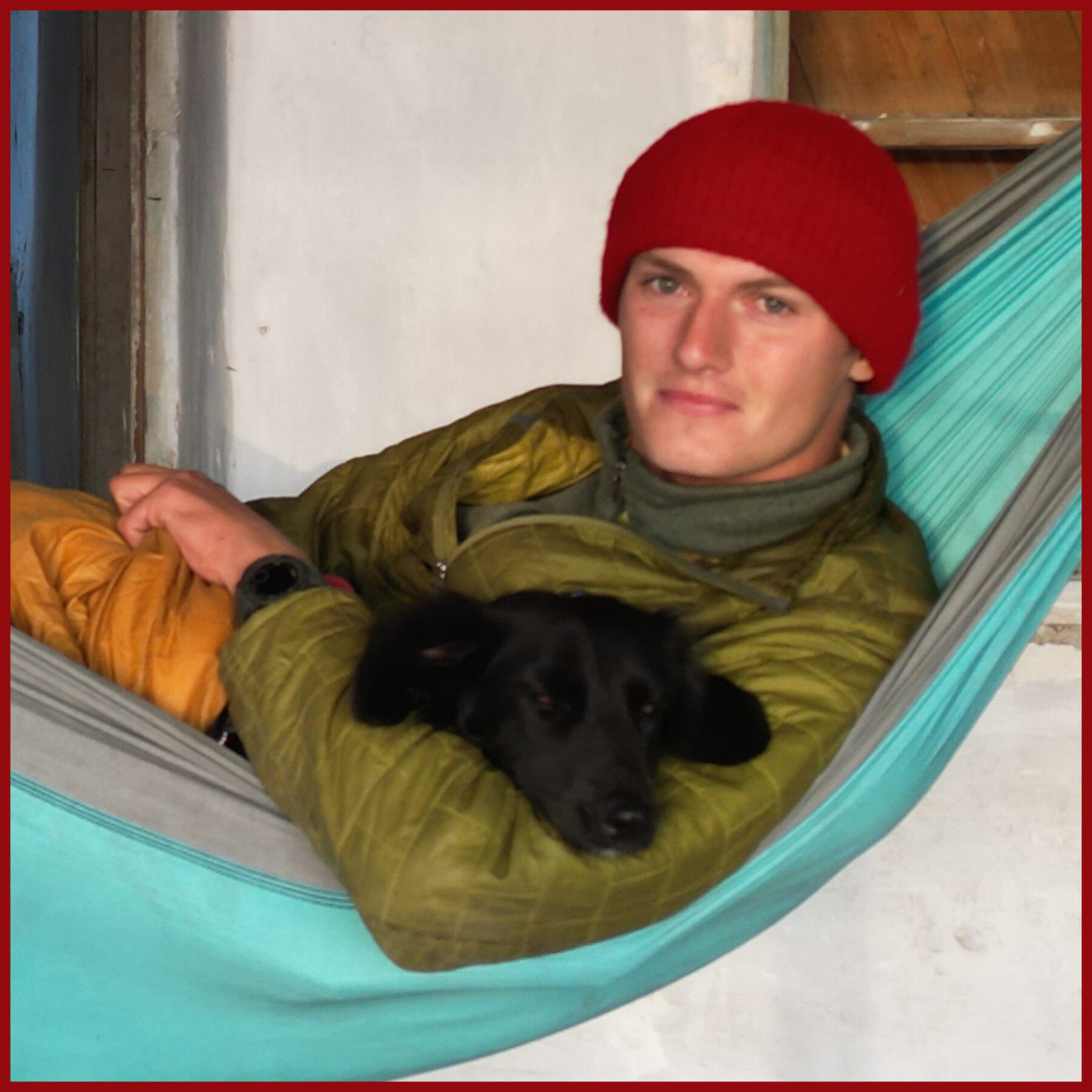 Portrait of Tassilo in a red wool hat lying in a teal hammock, cradling Fluffo the black dog asleep on his chest