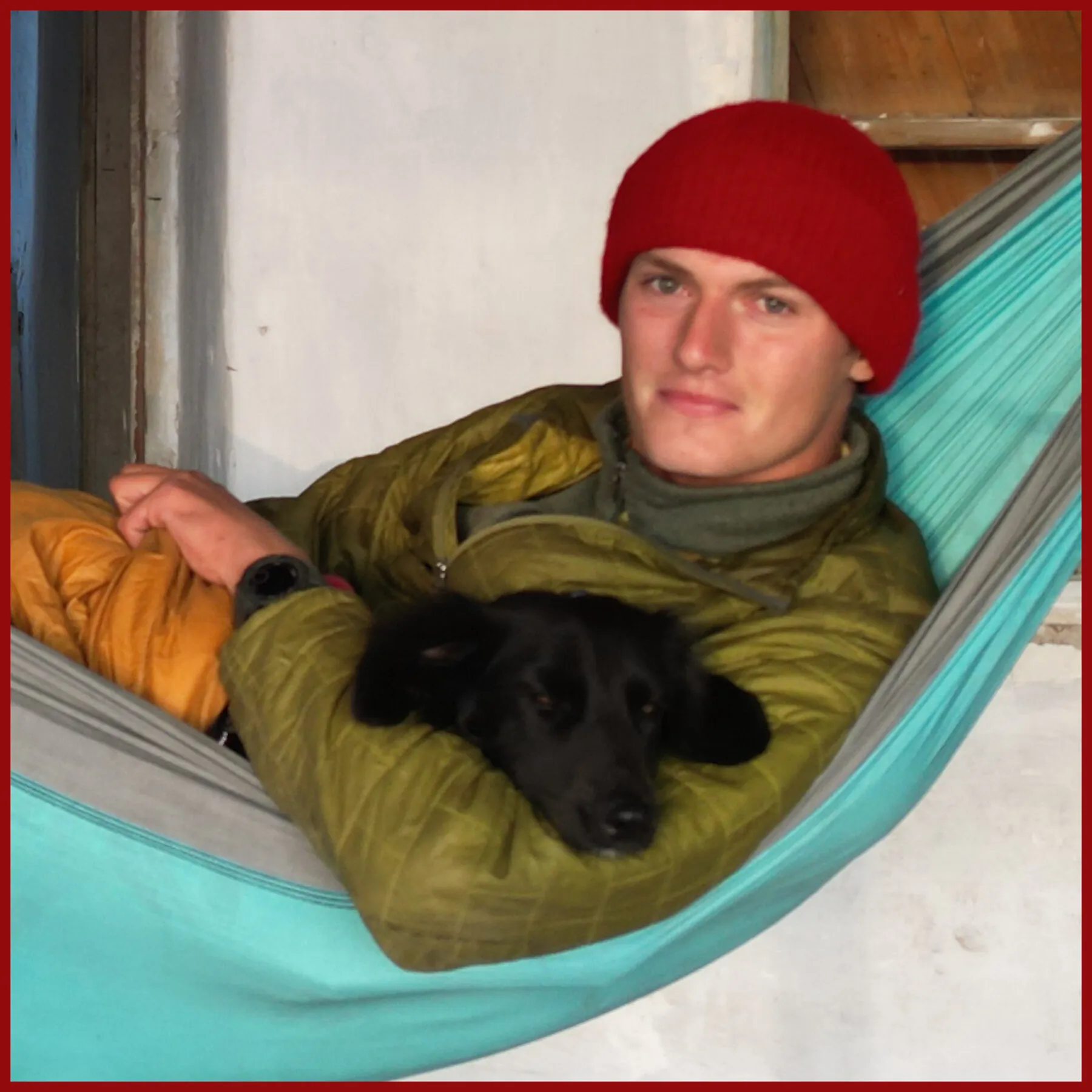 Portrait of Tassilo in a red wool hat lying in a teal hammock, cradling Fluffo the black dog asleep on his chest