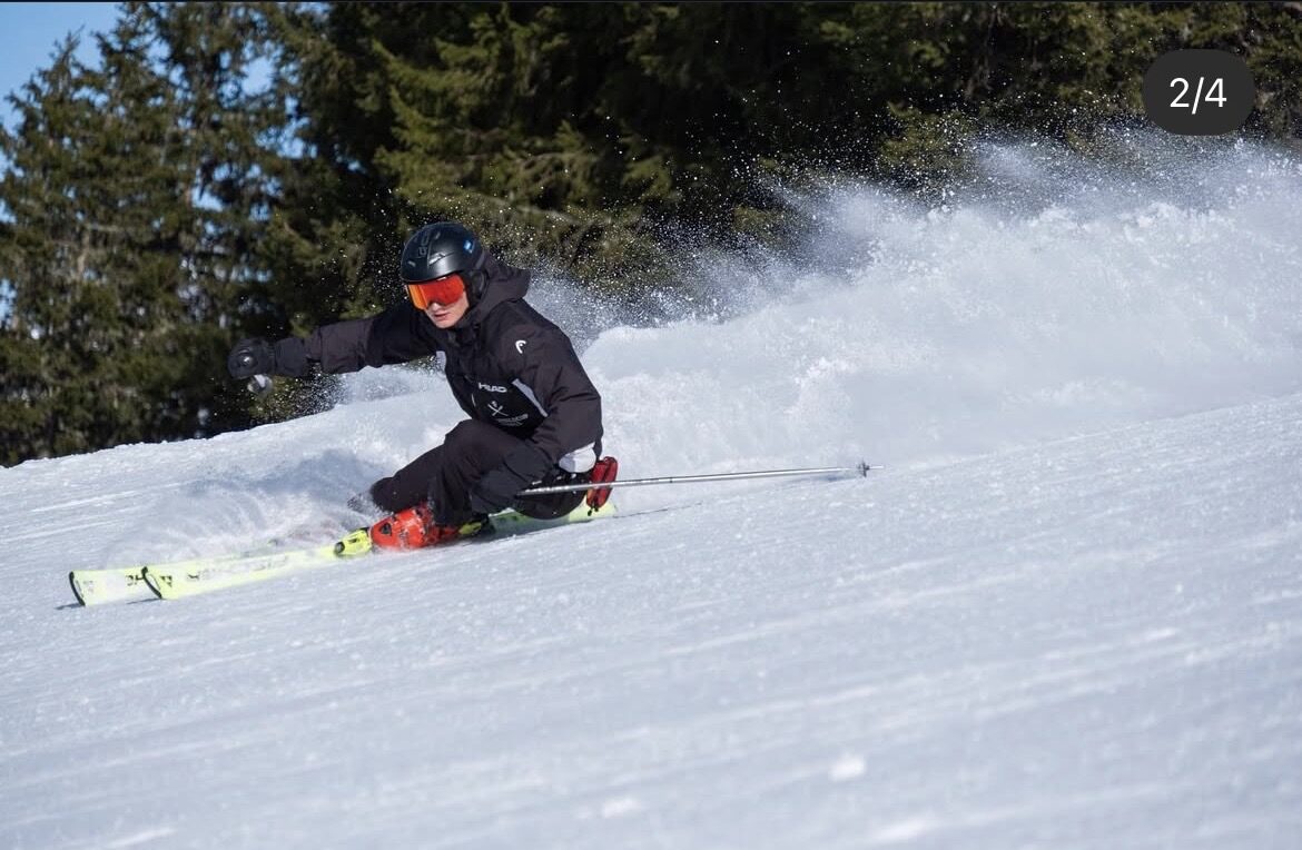 Tassilo skiing at speed, carving a deep turn with snow spray flying against dark pines