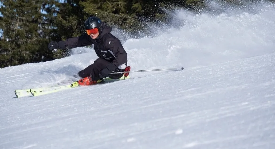 Tassilo skiing at speed, carving a deep turn with snow spray flying against dark pines