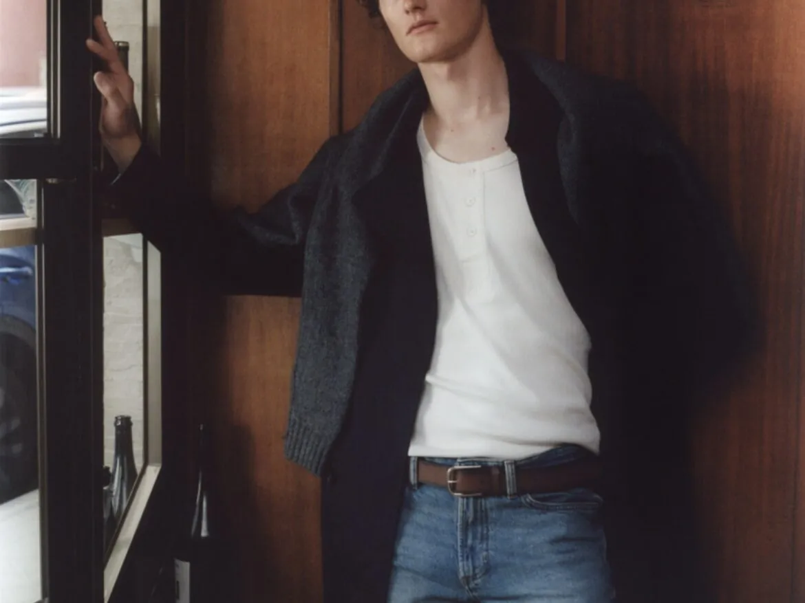 Portrait of Tassilo in a white tee and open dark shirt, leaning against a wood-panelled doorway