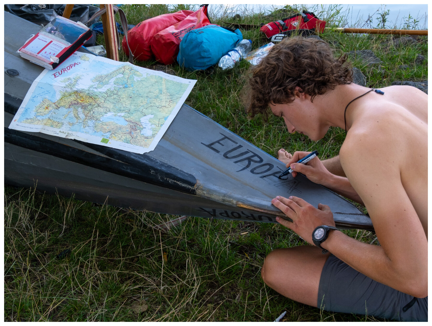 Tassilo kneeling on the grass, hand-painting the name EUROPA on the bow of his folding kayak, a marked-up map of Europe spread on the hull beside him