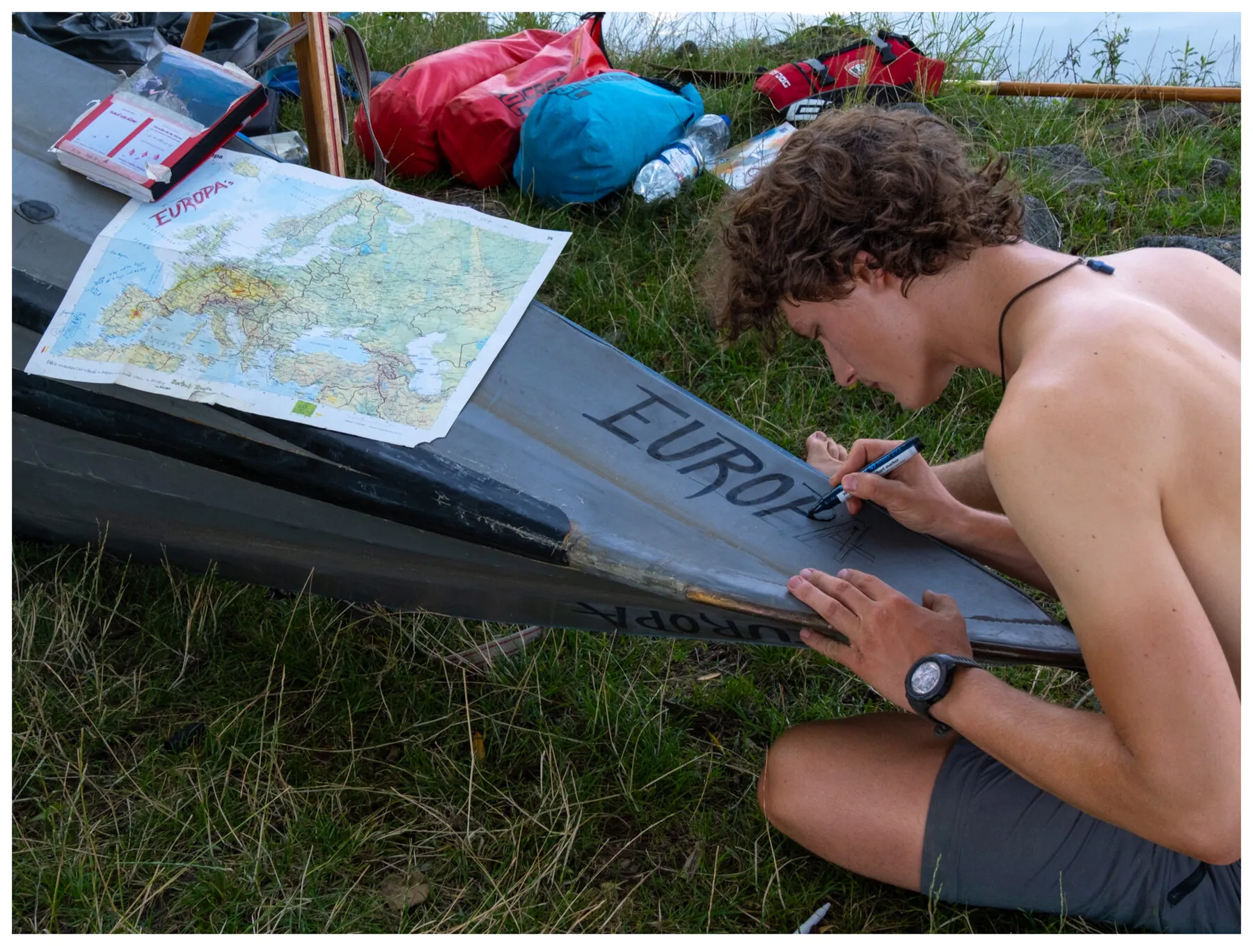 Tassilo kneeling on the grass, hand-painting the name EUROPA on the bow of his folding kayak, a marked-up map of Europe spread on the hull beside him