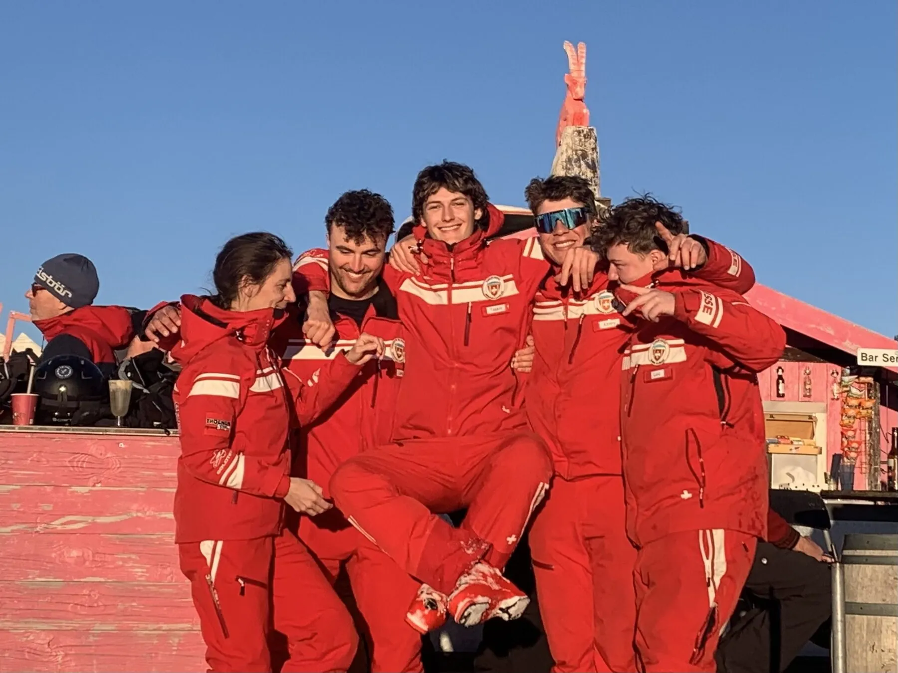 Tassilo laughing at the centre of a group of ski instructors in red uniforms, lifted off his feet at a sunny mountain bar