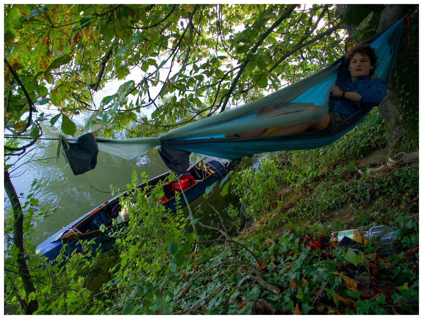 A man reading in a hammock strung over trees, a loaded kayak on the bank