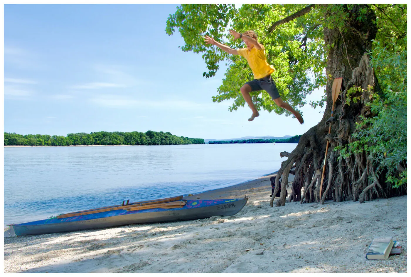 A young man caught mid-air leaping by a big riverside tree, kayak and dog on the beach
